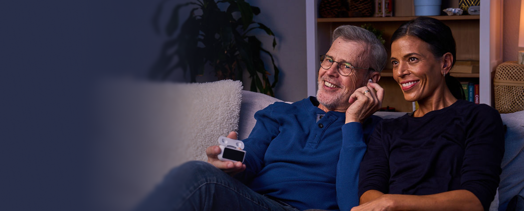 Man and woman sitting on a couch, smiling and holding a remote control.