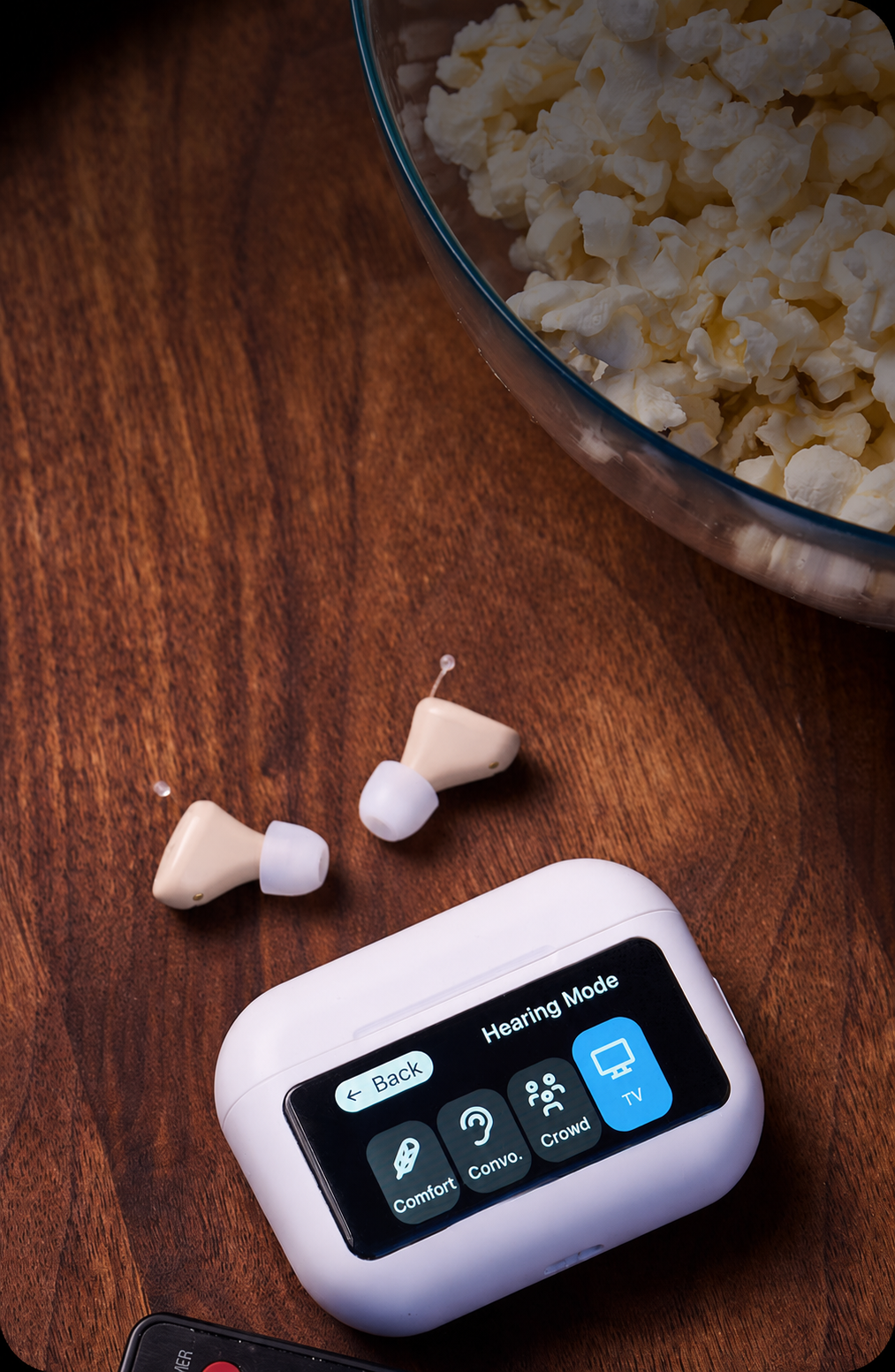 A pair of beige and white wireless hearing aids with their white charging case on a dark wooden table next to a bowl of popcorn.